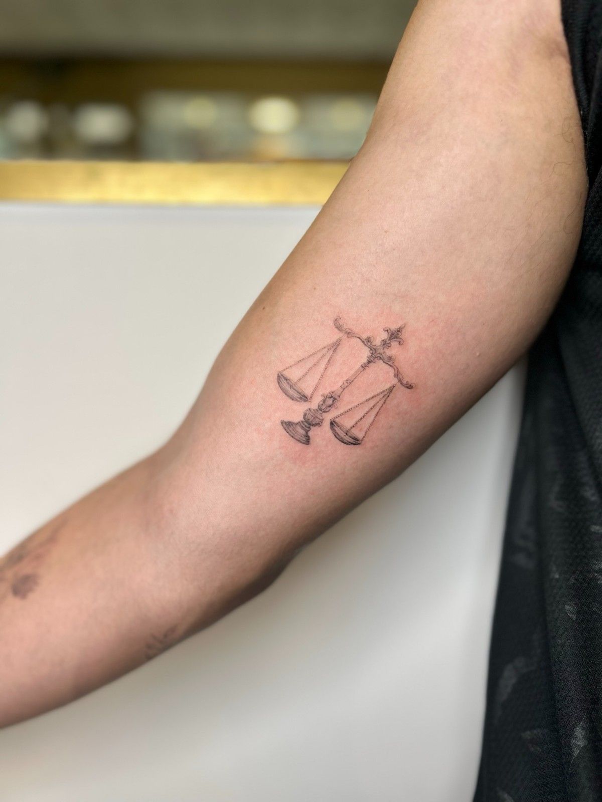 Tatuaje de balanza en el brazo, representando justicia o equilibrio, en tinta negra.