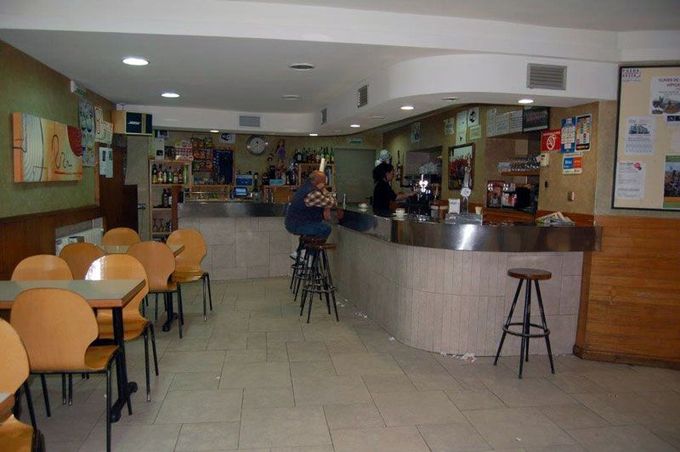 Cafetería espaciosa con mesas y taburetes, una barra curva y una persona de pie en el centro.