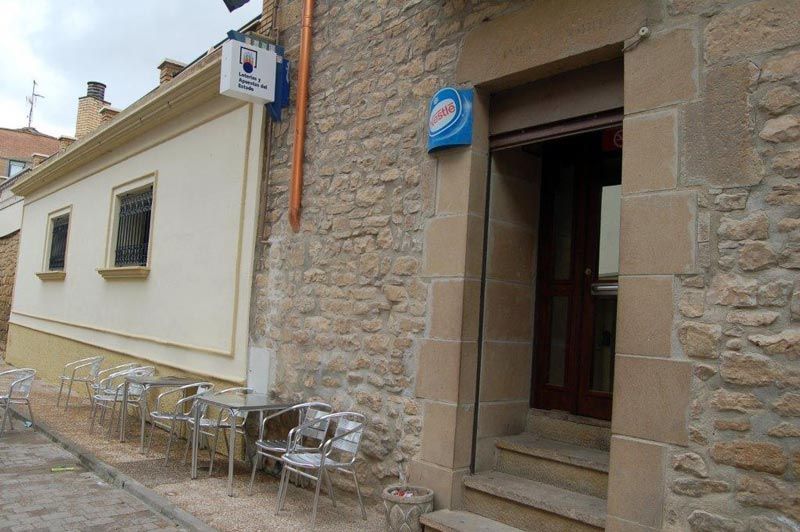 Edificio de piedra con una puerta de entrada y mesas y sillas de café al aire libre en una calle estrecha.