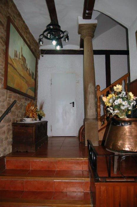 Un pasillo estrecho con escalones rojos, puerta blanca, columnas de piedra, lámpara colgante y flores en las mesitas auxiliares.