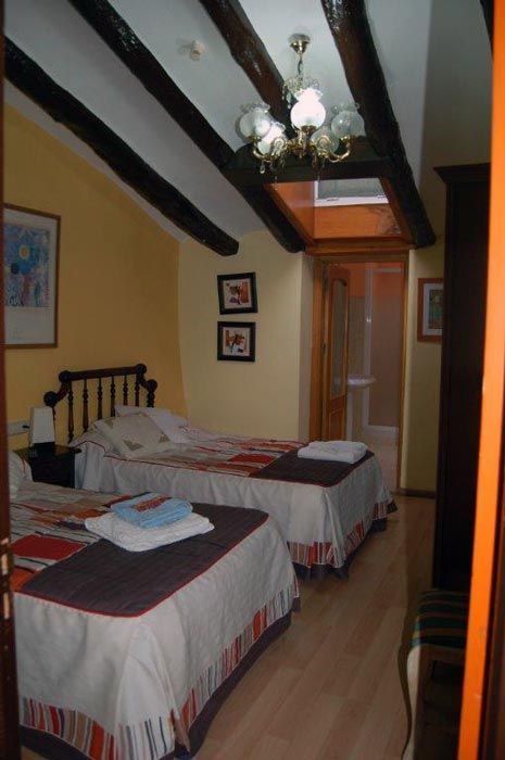 Habitación pequeña con dos camas individuales, vigas oscuras en el techo, lámpara de araña y una puerta con paredes de color amarillo cálido.