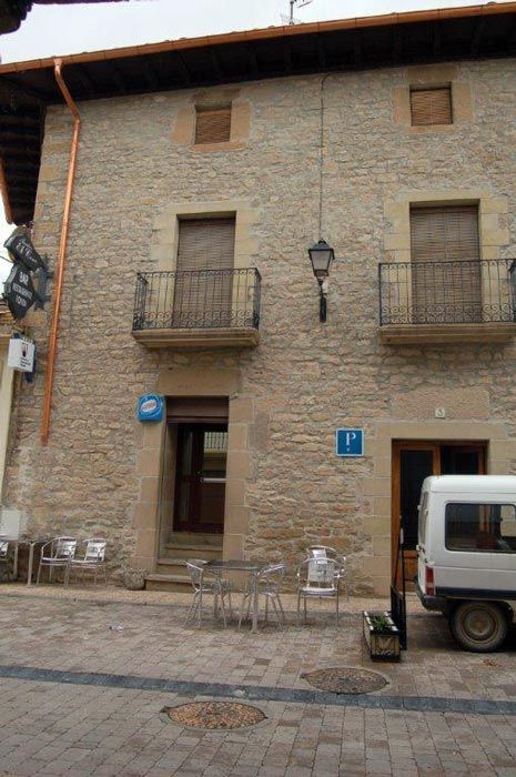 Fachada de un edificio de piedra con pequeños balcones, farolas, mesas de café y una furgoneta aparcada en una calle empedrada.