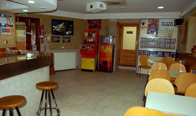 Interior de bar vacío con taburetes, mesas, máquinas recreativas e iluminación cálida.