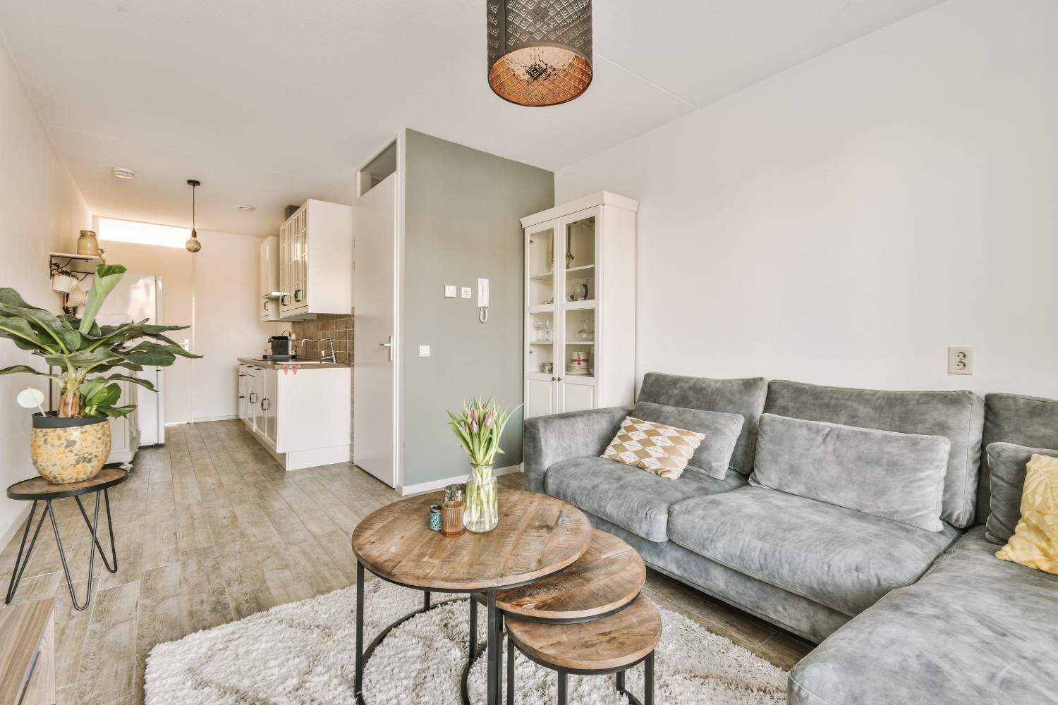 Una acogedora sala de estar con un sofá gris, mesas de centro de madera apiladas, una vitrina blanca y una cocina al fondo.