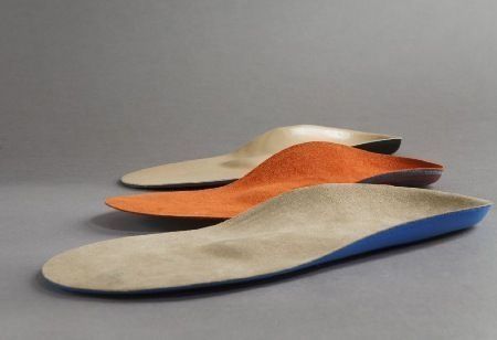 Drei individuell angefertigte orthopädische Einlegesohlen in Beige-, Orange- und Blautönen, angeordnet.
