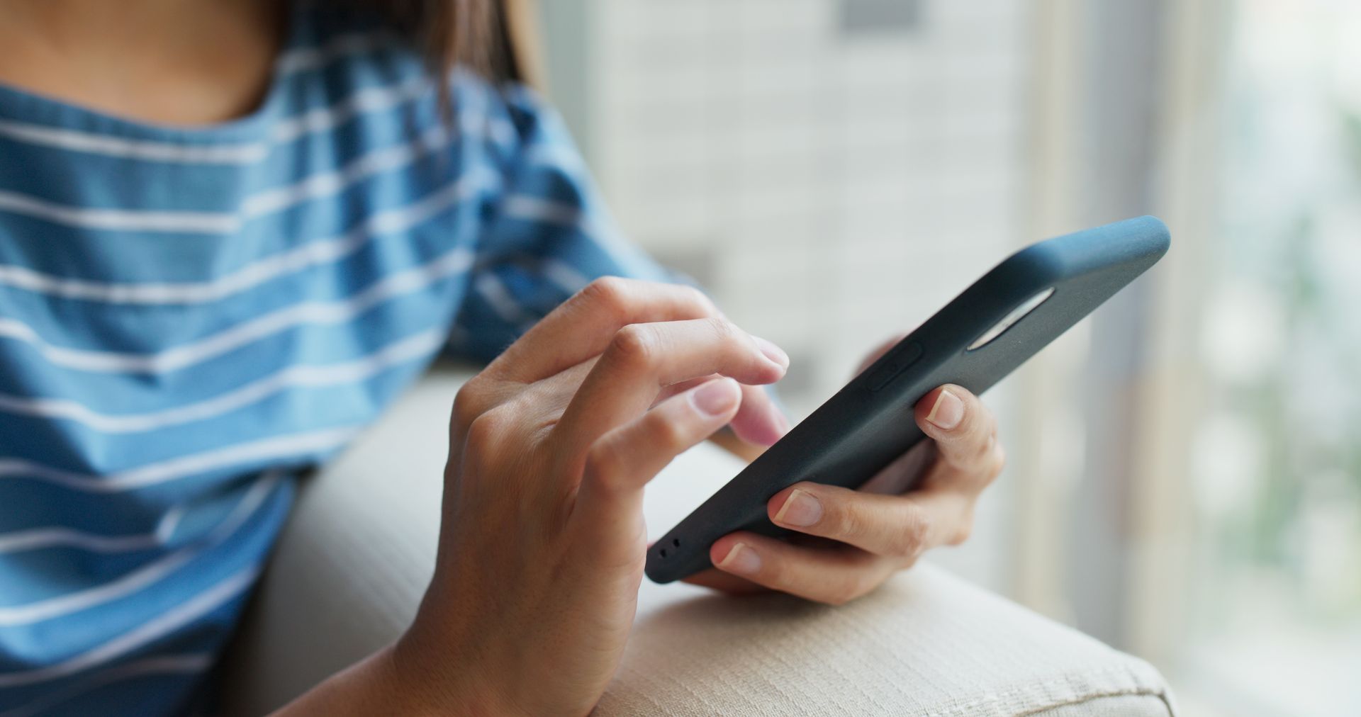 Une personne utilise un smartphone sur un canapé, vêtue d'une chemise à rayures bleues.