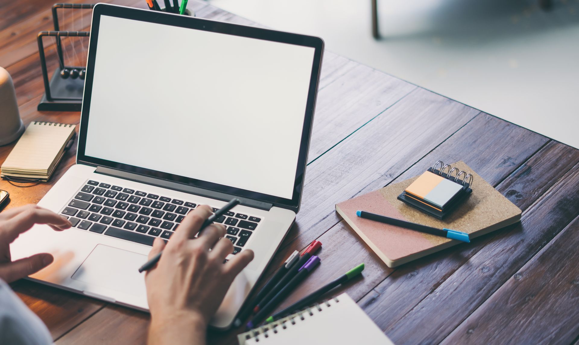 Des mains tapent sur un ordinateur portable à un bureau en bois, avec des cahiers et un stylo à proximité.