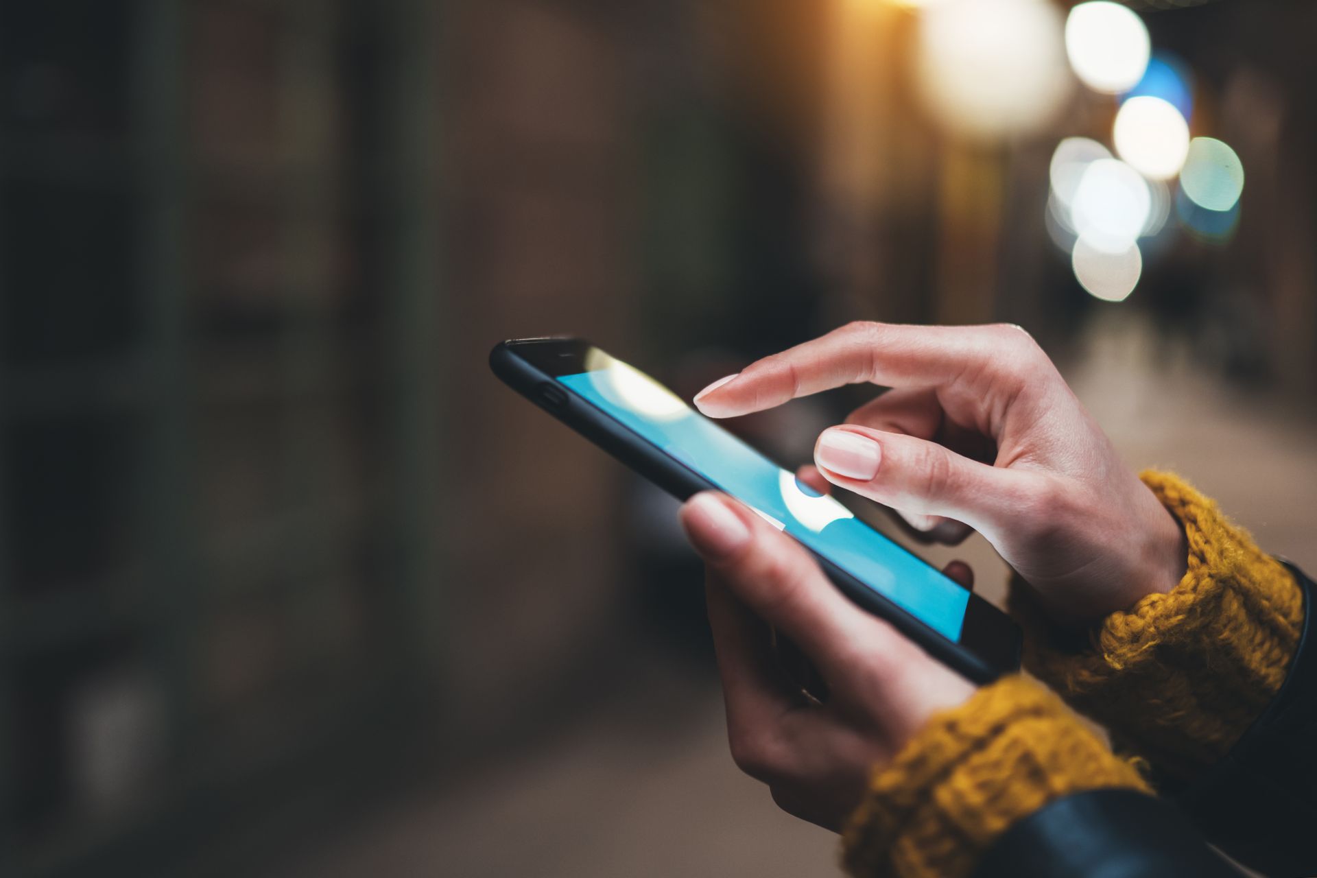 Des mains utilisent un smartphone à écran bleu en extérieur, la nuit, avec des lumières floues en arrière-plan.