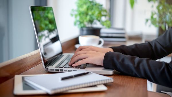 Une personne tape sur un ordinateur portable à un bureau en bois, avec un carnet et une tasse de café à proximité.