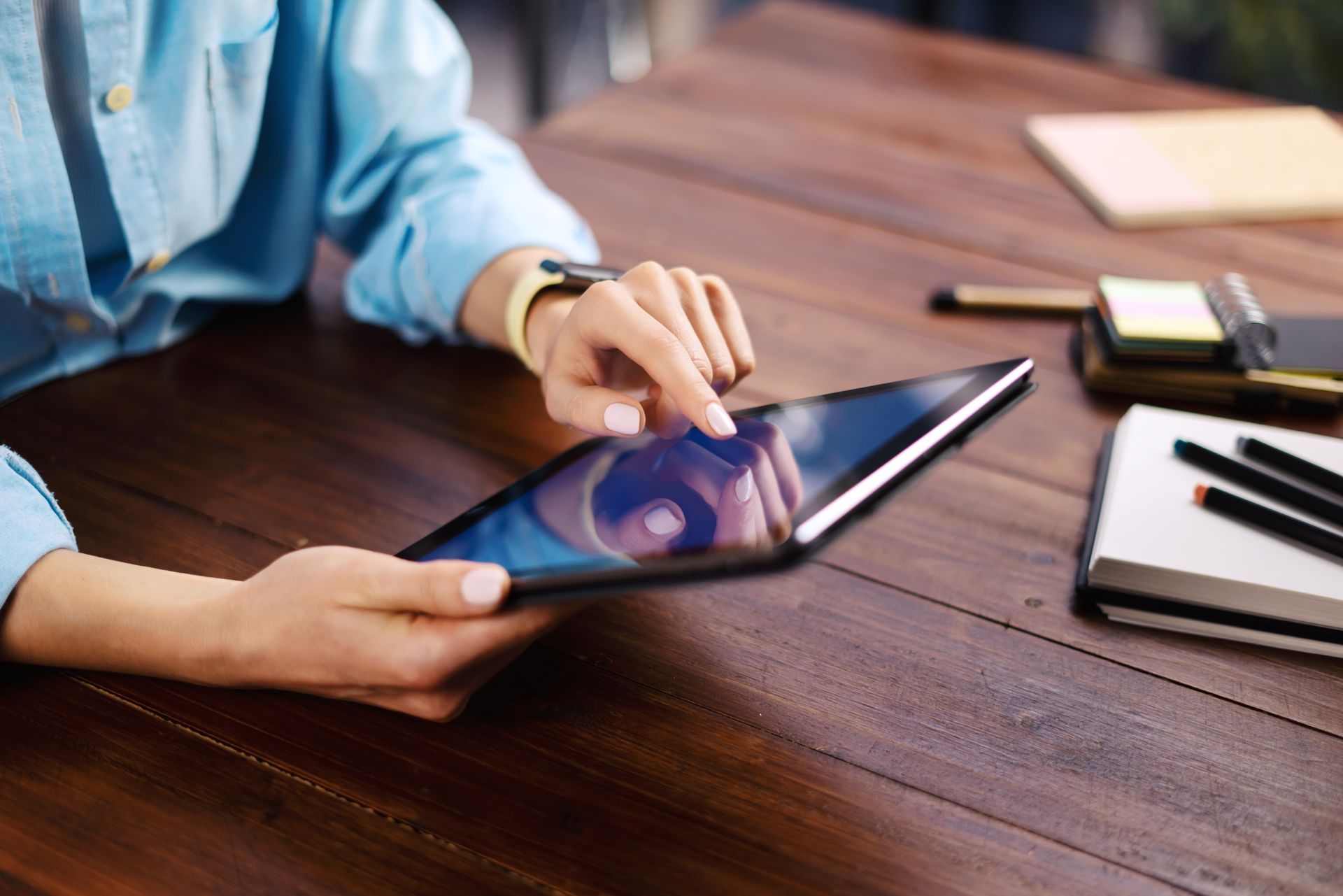 Une personne utilise une tablette à un bureau en bois, avec un ordinateur portable et des lunettes à proximité.
