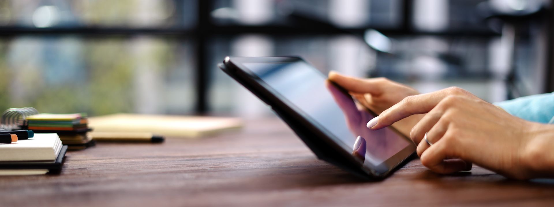 Des mains utilisent une tablette sur un bureau en bois, avec des cahiers et une tasse de café à proximité.