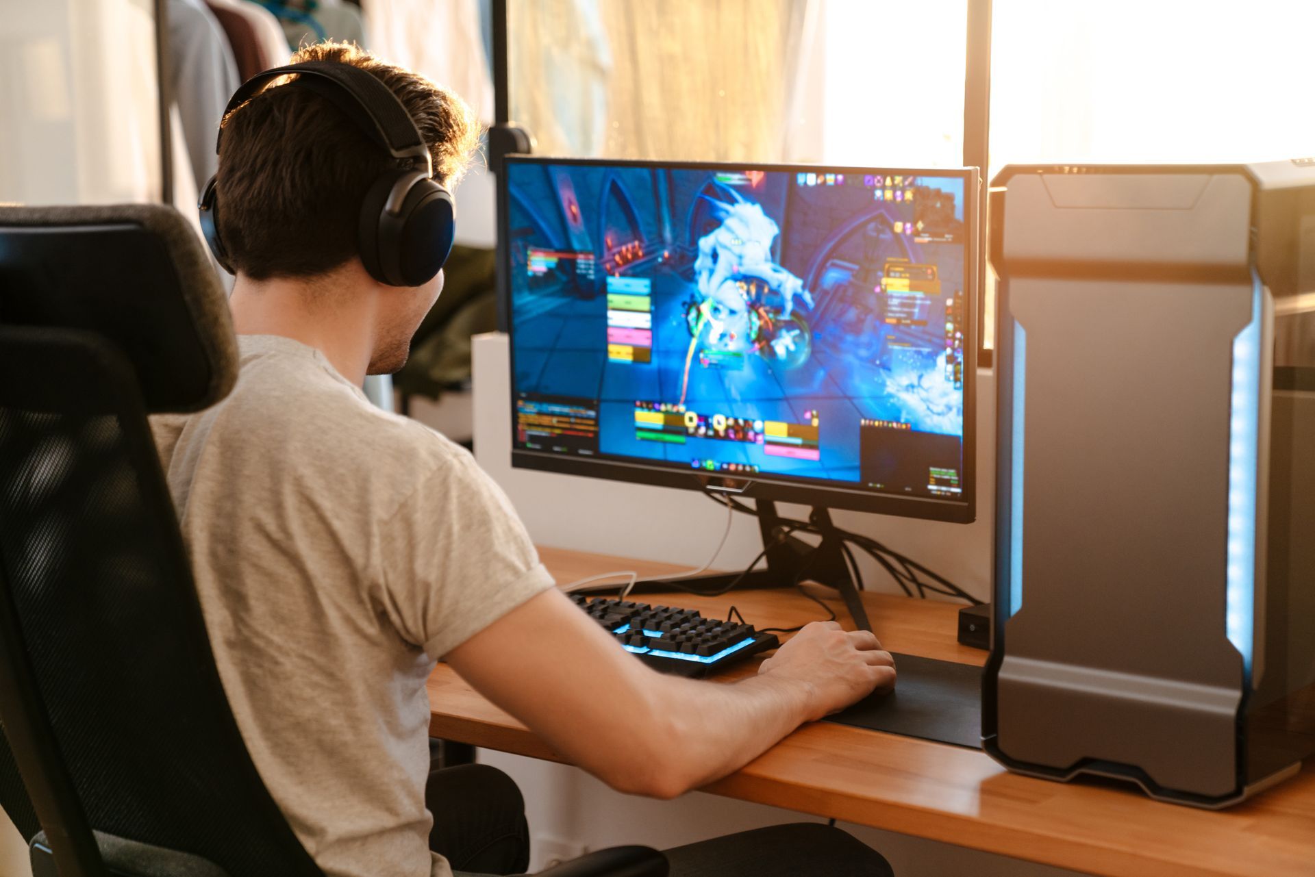Une personne joue sur un ordinateur de bureau avec un casque audio dans une pièce ensoleillée.