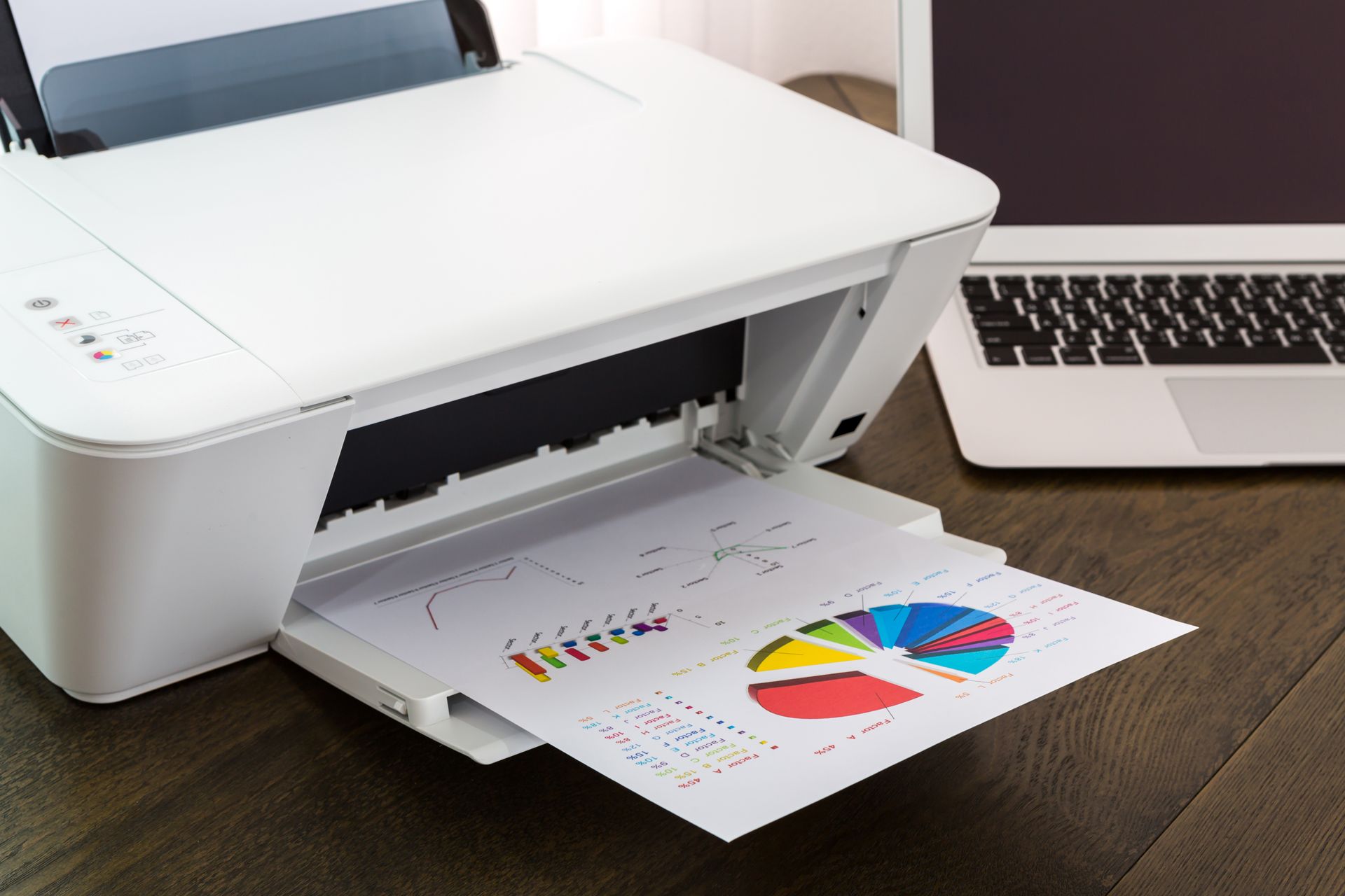 Une imprimante blanche imprime un graphique circulaire en couleur sur du papier à côté d'un ordinateur portable posé sur un bureau.