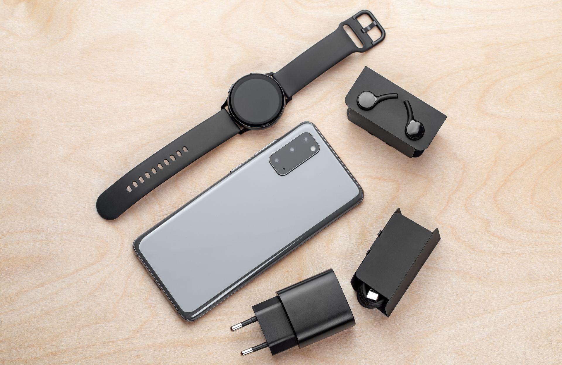 Montre connectée noire, téléphone argenté, écouteurs et chargeur disposés sur une surface en bois clair.