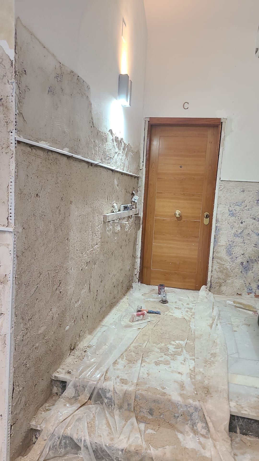 Baño en construcción con paredes parcialmente alicatadas, puerta y aplique de pared.