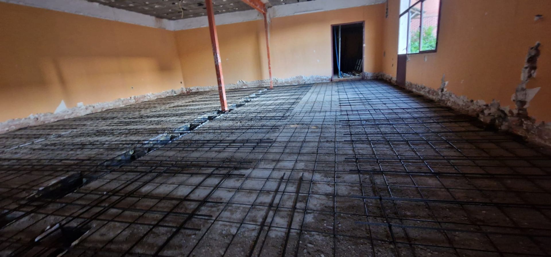 Vista interior de una habitación en construcción, con rejilla de armadura expuesta en el piso y paredes de color naranja.