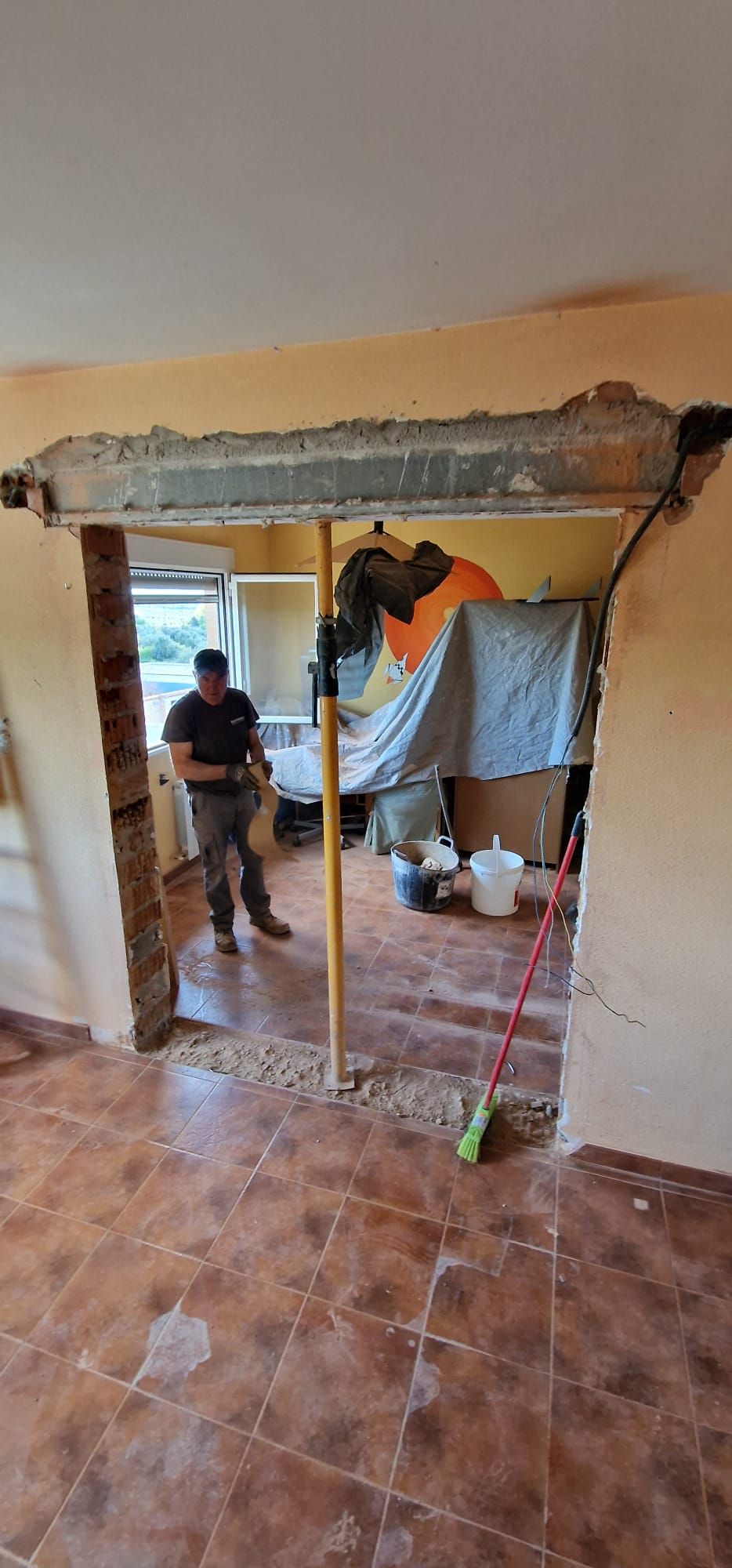 Hombres demoliendo una puerta en una habitación con piso de baldosas marrones; paredes amarillas, herramientas y escombros visibles.