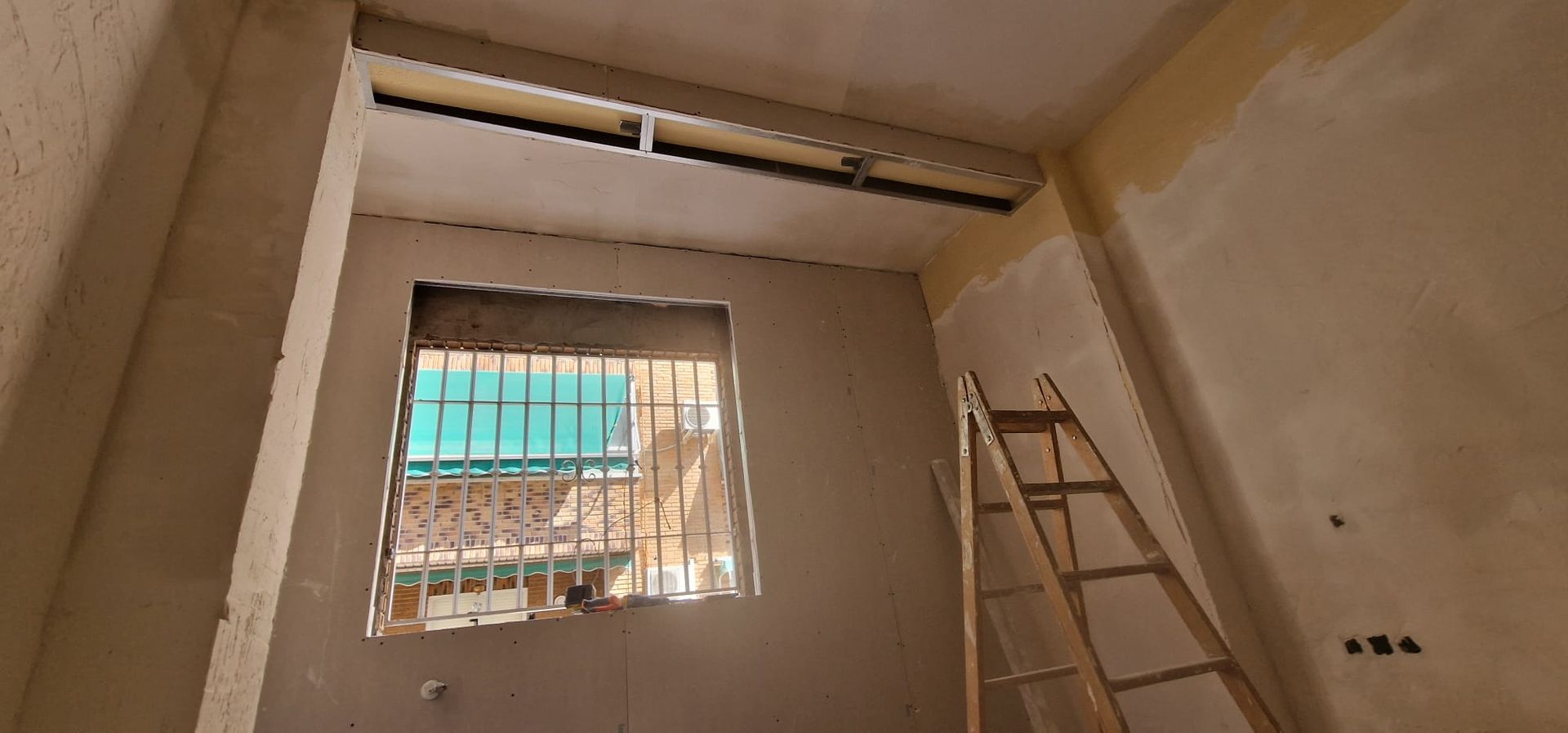 Vista interior de una habitación en reforma. Una escalera está apoyada contra la pared, junto a una ventana con rejas.