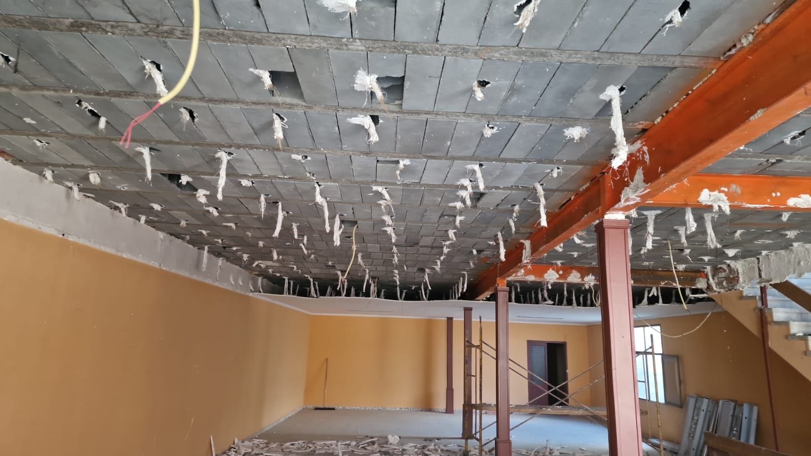 Vista interior de un edificio en construcción. Techo dañado con vigas expuestas, pared naranja y escalera.