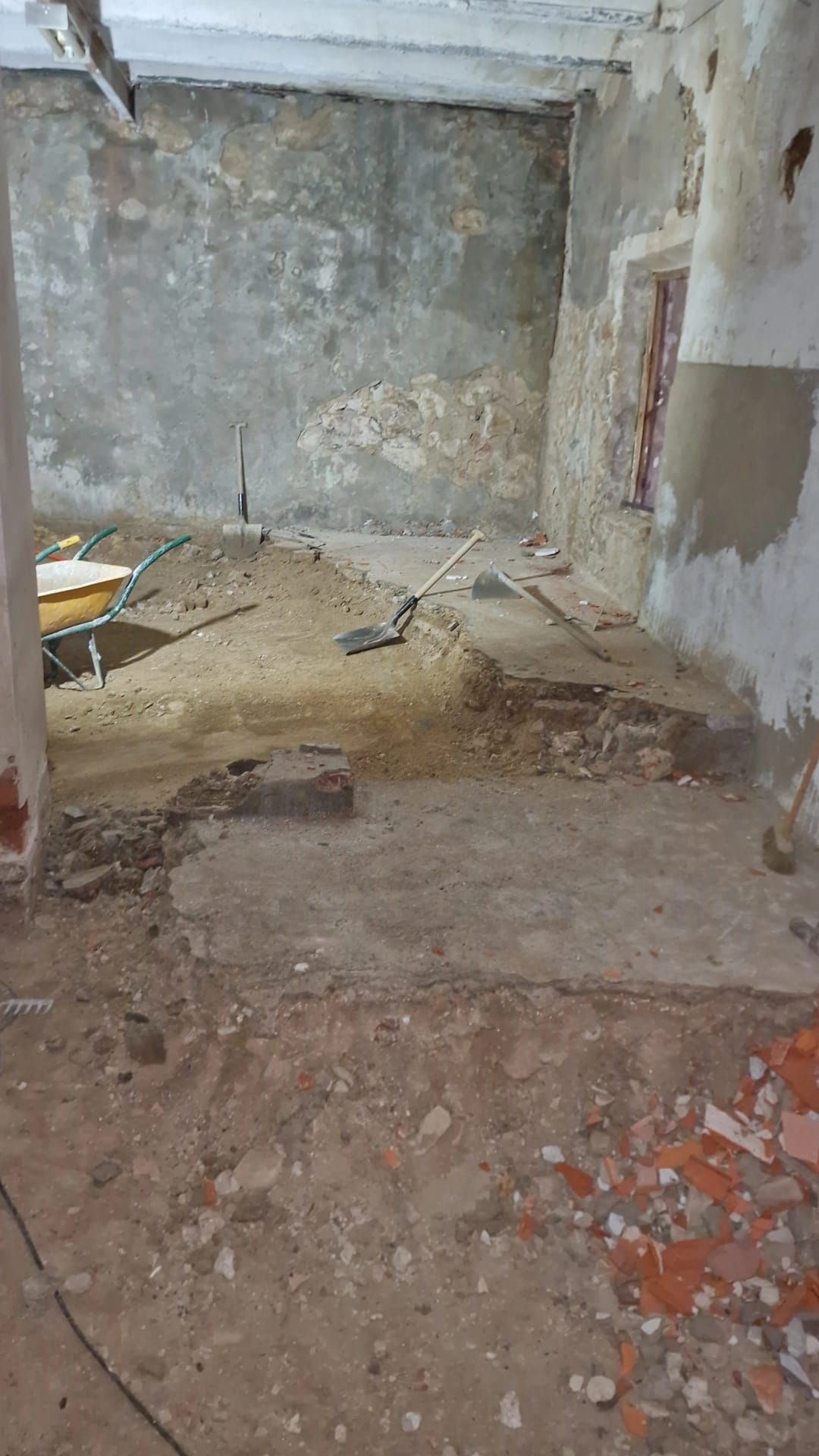 Demolición interior de una habitación con paredes y suelo a la vista. Escombros y herramientas esparcidos.