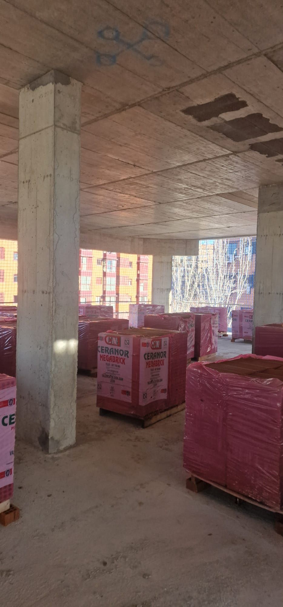 Interior de un edificio en construcción, con pilas de materiales rosas sobre palés. Pilares y techo de hormigón gris.