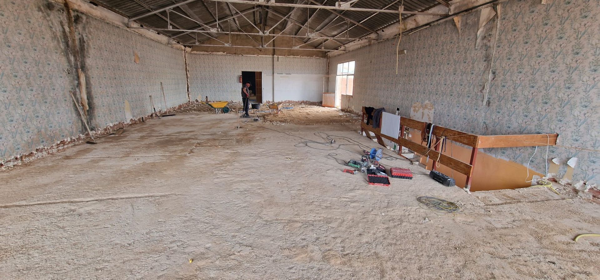 Dentro de un edificio ruinoso con suelo de tierra. Una persona está de pie cerca de una puerta. Hay escombros y muebles esparcidos por todas partes.