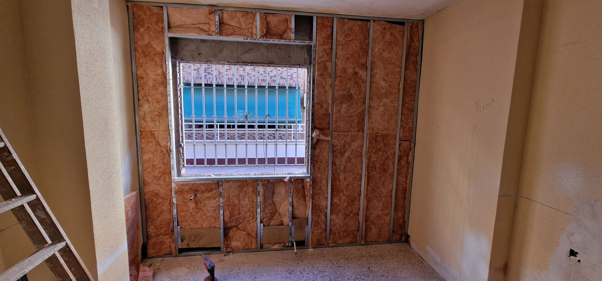 Interior de la habitación en construcción con ventana y rejas de seguridad. Aislamiento y montantes metálicos visibles.
