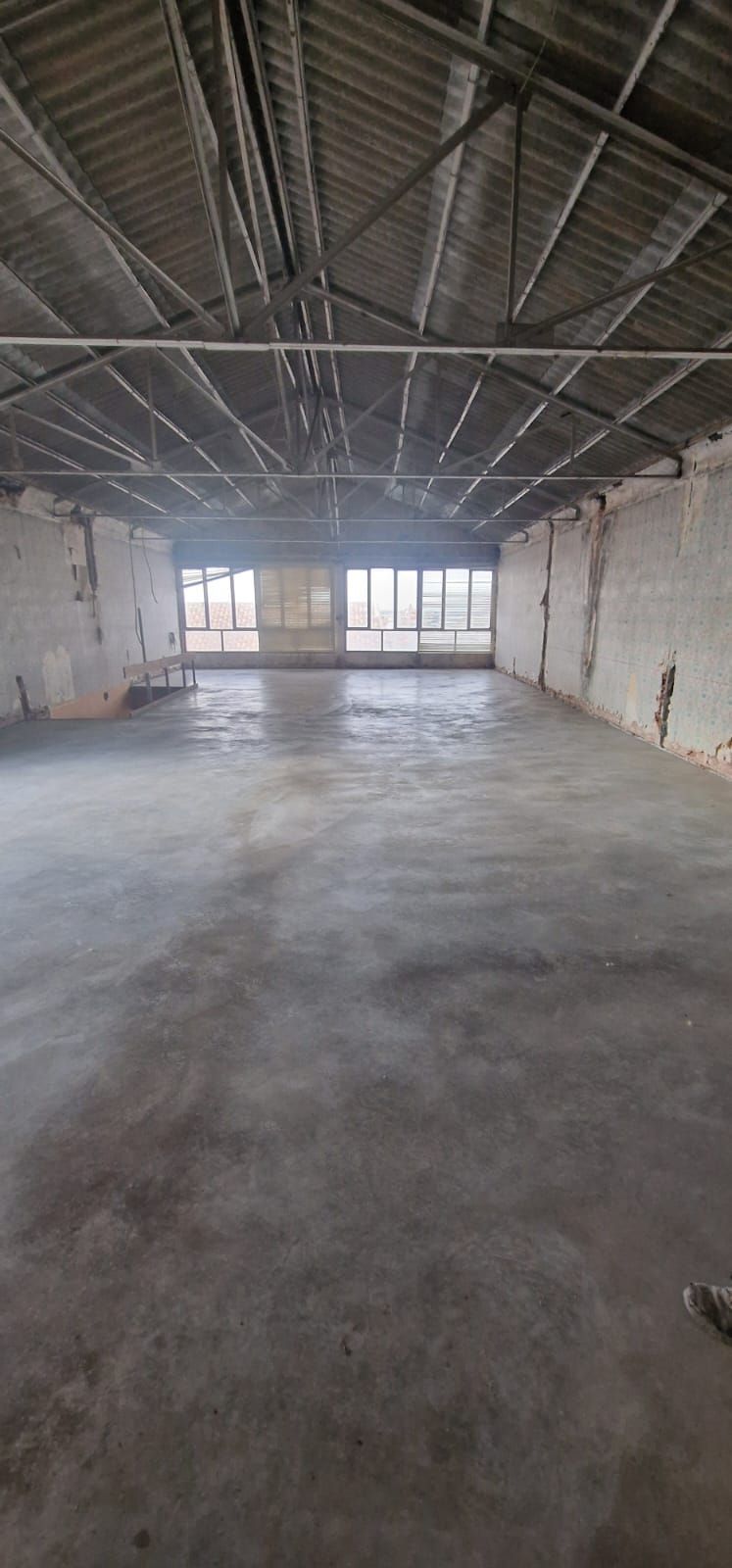 Interior vacío y desgastado de un gran edificio industrial con techos altos y piso de concreto.