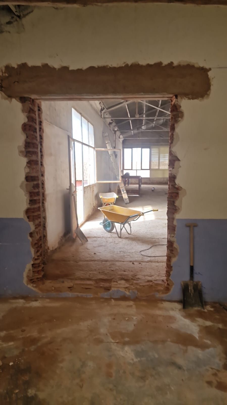 Vista interior de una puerta cortada a través de una pared de ladrillos, que conduce a un sitio de construcción con herramientas y una carretilla.