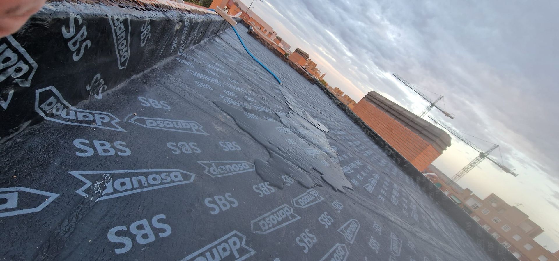 Material de techado negro en un día nublado, con algunas grietas visibles. Edificios a lo lejos.