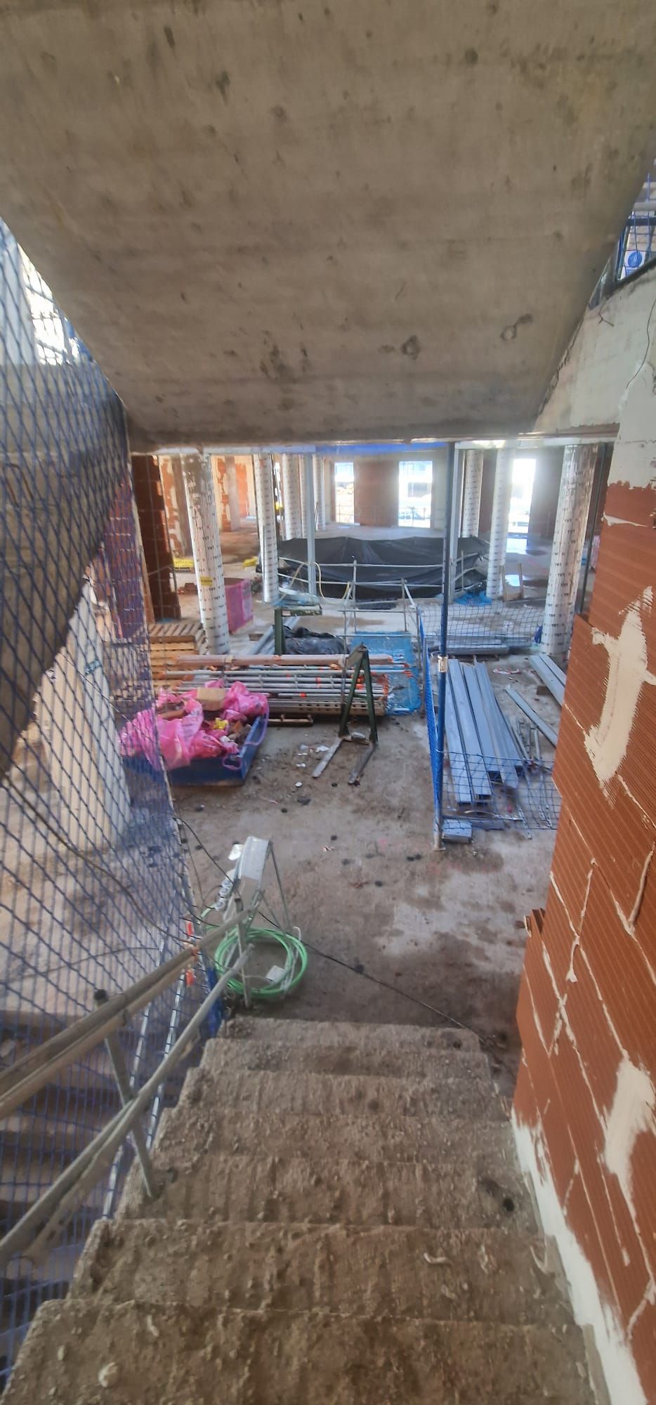 Interior de una obra en construcción. Unas escaleras de hormigón conducen a una sala con materiales de construcción, paredes de ladrillo y puertas abiertas.
