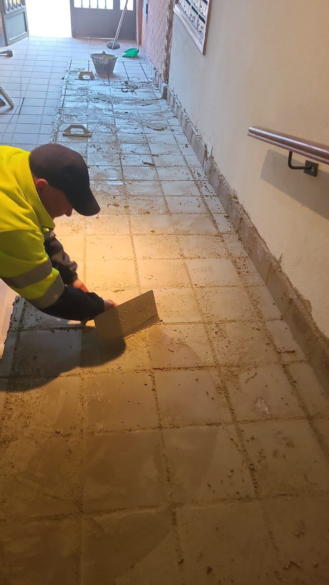 Persona con chaleco reflectante aplicando mortero con llana sobre las baldosas del pasillo.