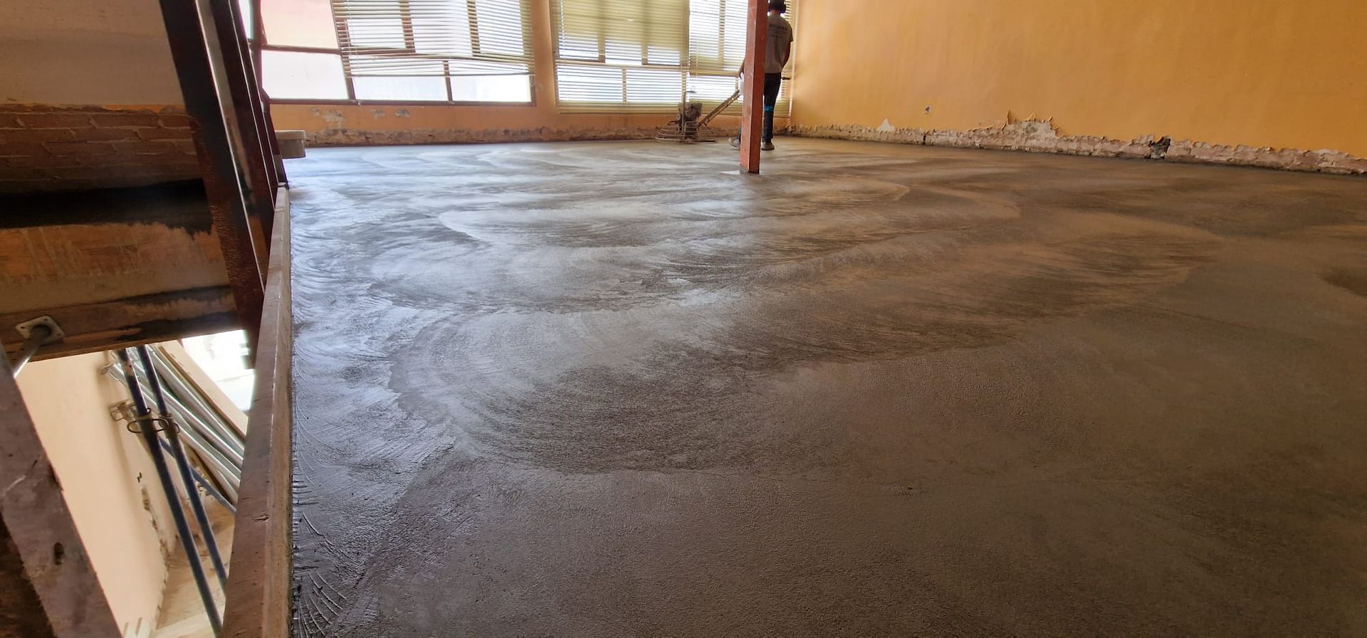 Piso de hormigón recién vertido en el interior de un edificio, con escalera expuesta a la izquierda y ventanas al fondo.