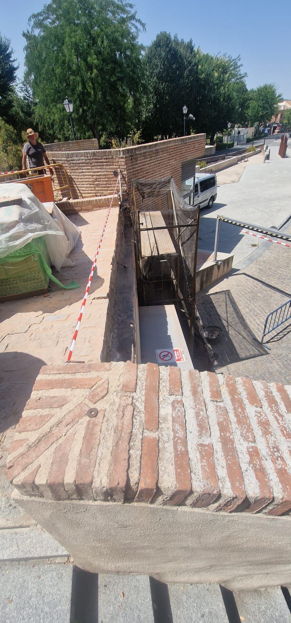 Obra con estructuras de ladrillo y hormigón. Un trabajador está cerca de los materiales, rodeado de árboles y una calle.