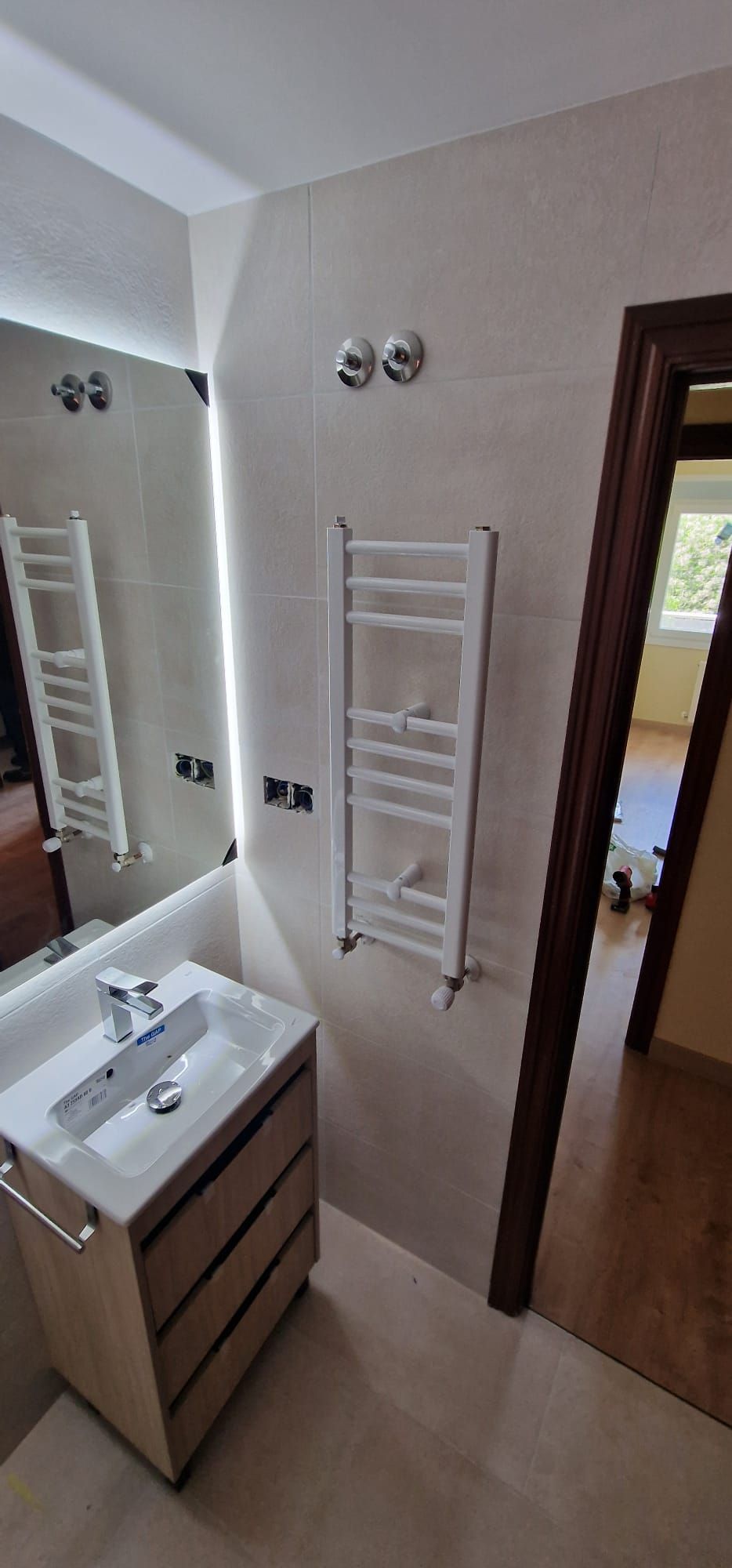 Baño con lavabo, espejo y toallero eléctrico. La puerta da a una habitación con suelo de madera.