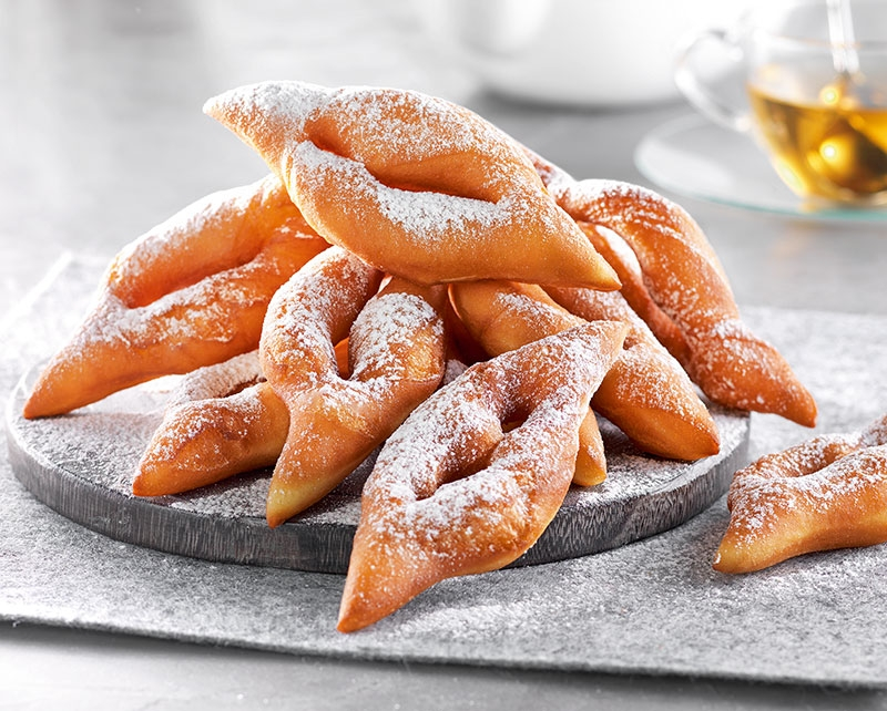 Des beignets frits saupoudrés de sucre glace, empilés sur une dalle de pierre grise.