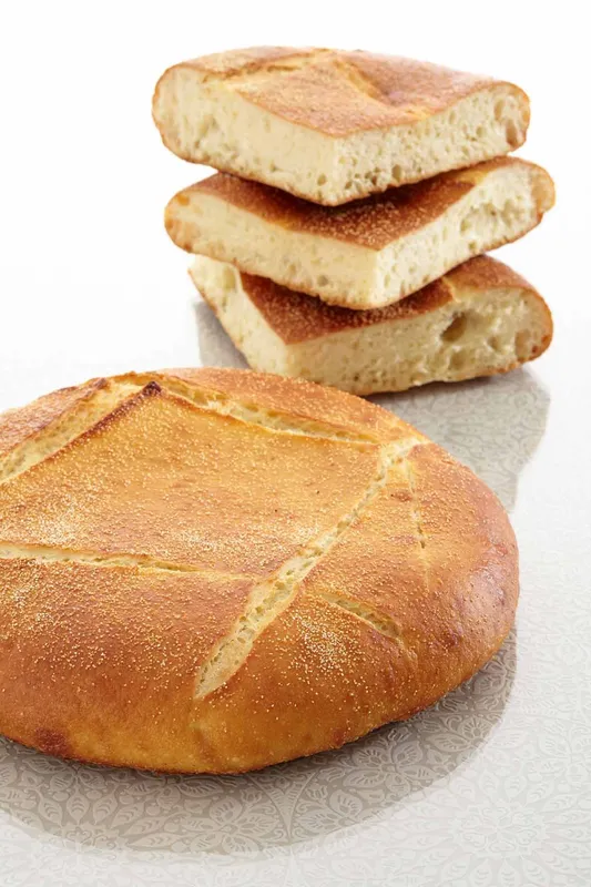 Un pain rond et doré, orné d'entailles en forme d'étoile sur le dessus. Des tranches sont empilées derrière.