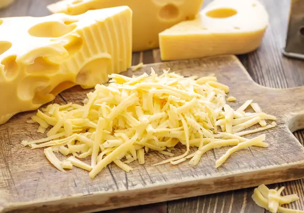 Fromage jaune râpé sur une planche en bois, avec des morceaux de fromage suisse en arrière-plan.
