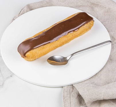 Éclair au chocolat sur une assiette blanche, accompagné d'une cuillère en argent, le tout posé sur une nappe en lin.