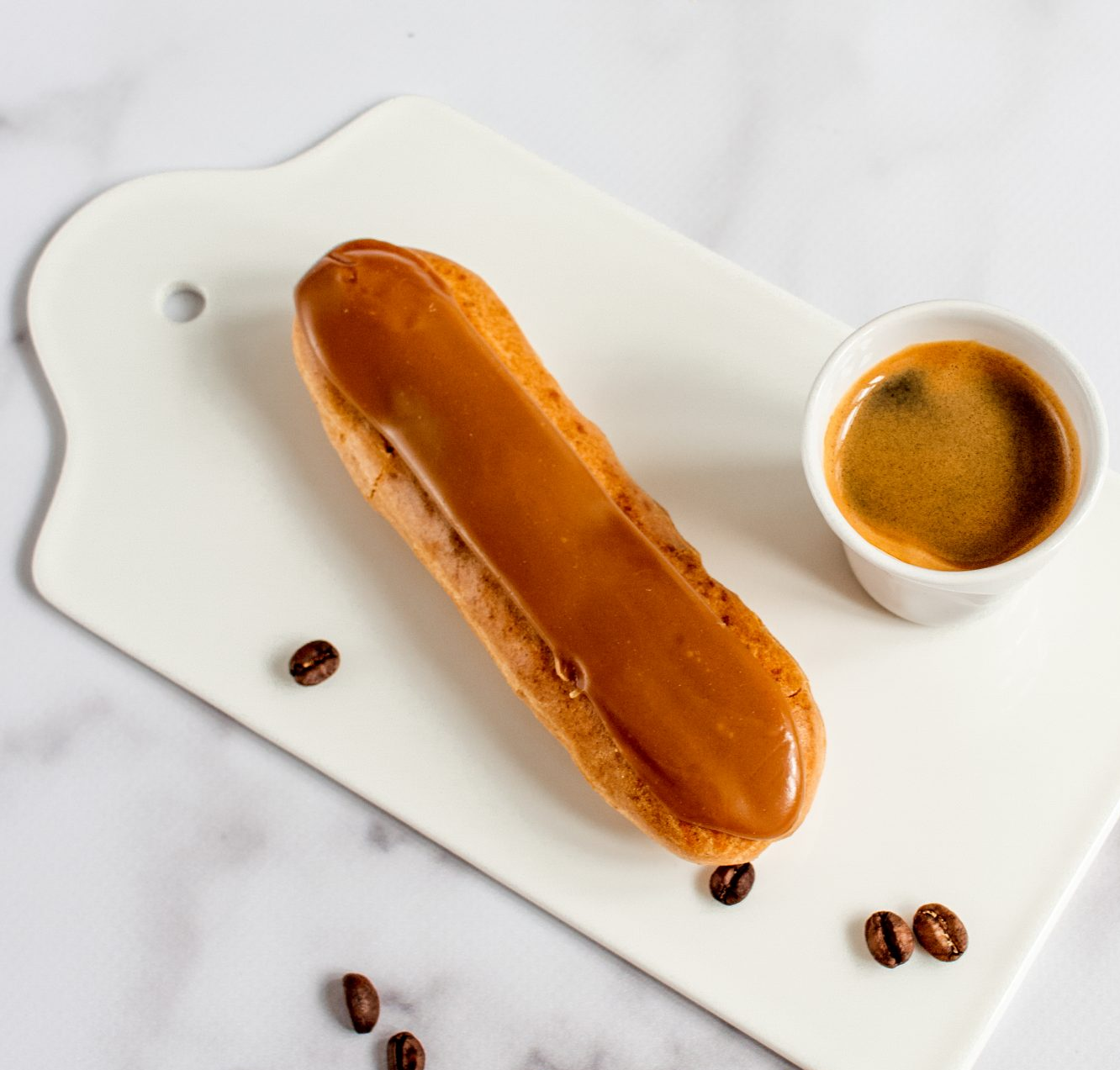 Éclair nappé de caramel à côté d'un expresso sur une planche à découper blanche, avec des grains de café.
