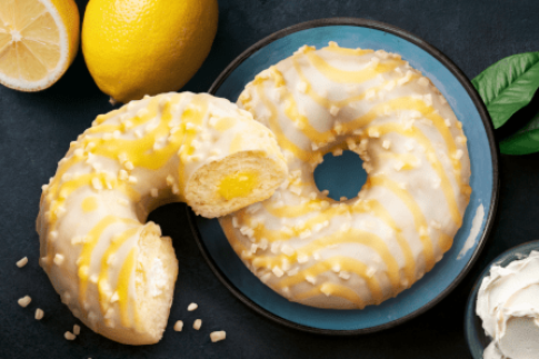 Beignet fourré au citron avec glaçage blanc et jaune, noix hachées, demi-citron sur une assiette bleu foncé.