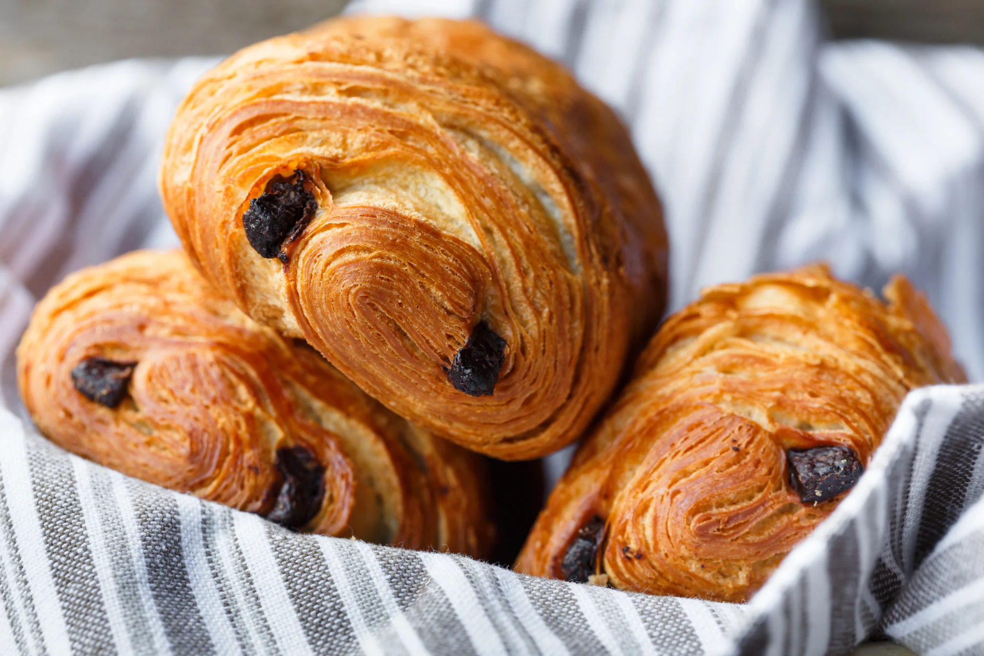 Trois pains au chocolat dorés et feuilletés, présentés dans un panier doublé de lin rayé.