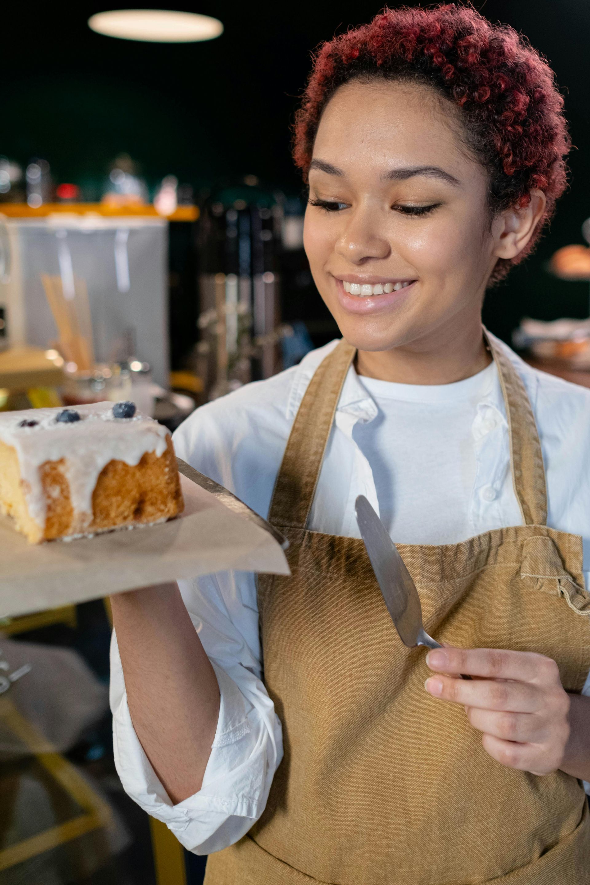 Une personne tenant une part de gâteau glacé aux myrtilles, souriante dans un café, portant un tablier.