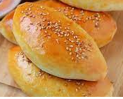 Pâtisseries ovales, dorées et parsemées de graines de sésame, empilées sur une surface claire.