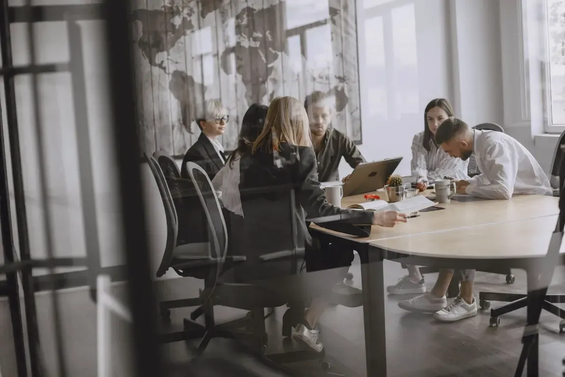 Reunión de equipo alrededor de una mesa en una luminosa sala de conferencias, con ordenadores portátiles y papeles esparcidos.