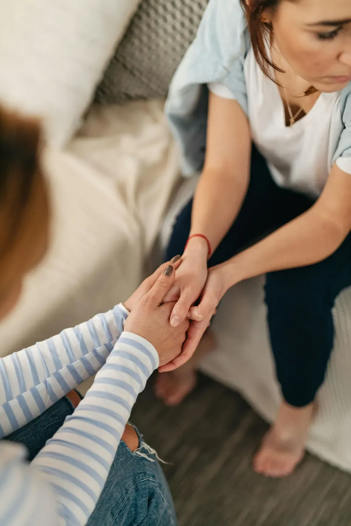 Una mujer sostiene suavemente las manos de otra persona mientras están sentadas en un sofá en el interior de una vivienda.