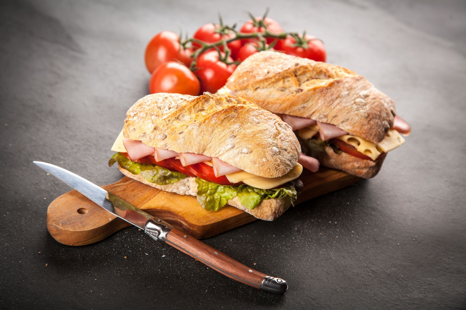Dos sándwiches están sobre una tabla de cortar de madera con tomates en el fondo.