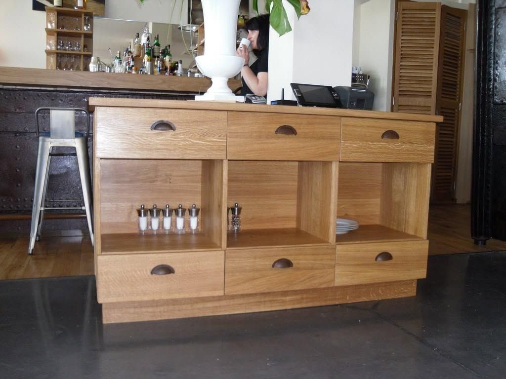 Îlot de cuisine en bois avec des tiroirs et des étagères ouvertes, accueillant des bouteilles et de petits objets dans une cuisine.