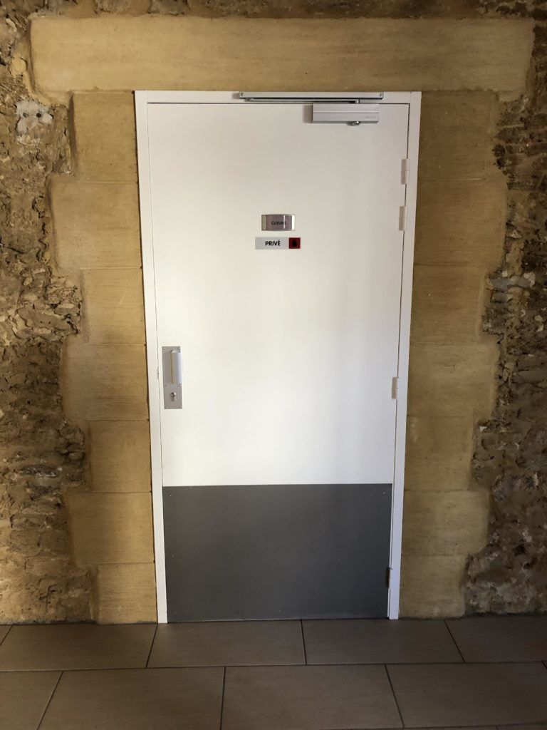 Porte blanche avec panneau inférieur gris, encastrée dans un mur de pierre dans un couloir.