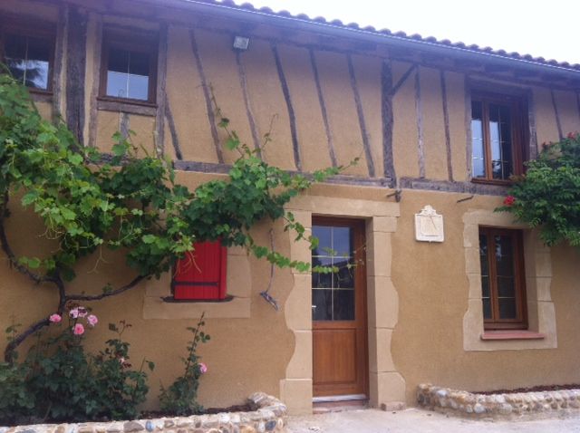 Maison rustique couleur beige, avec des poutres en bois, des plantes grimpantes vertes, des volets rouges et une porte d’entrée en bois